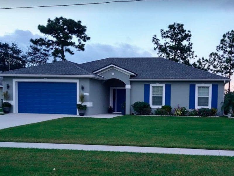 Modern gray house with blue garage door, lush garden, and tall pine trees in the background.